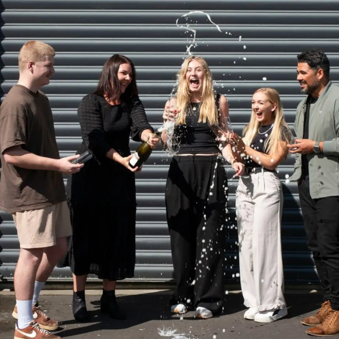 A dark green champagne bottle being opened by two people, with sparkling liquid spraying upward in a celebratory arc.