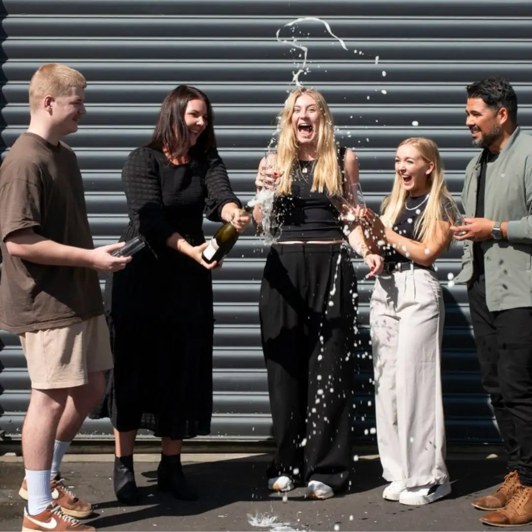 A dark green champagne bottle being opened by two people, with sparkling liquid spraying upward in a celebratory arc.