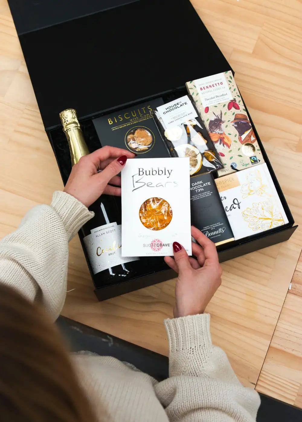A white rectangular box labeled ’bubbly bears’ with a circular window revealing golden-brown candy inside, held by hands with red nail polish.