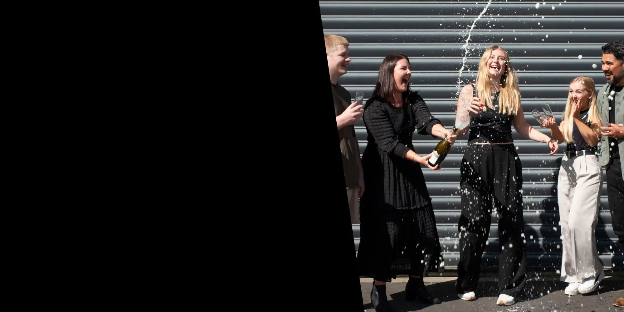 A woman in a black dress holds an open champagne bottle as it sprays sparkling liquid.