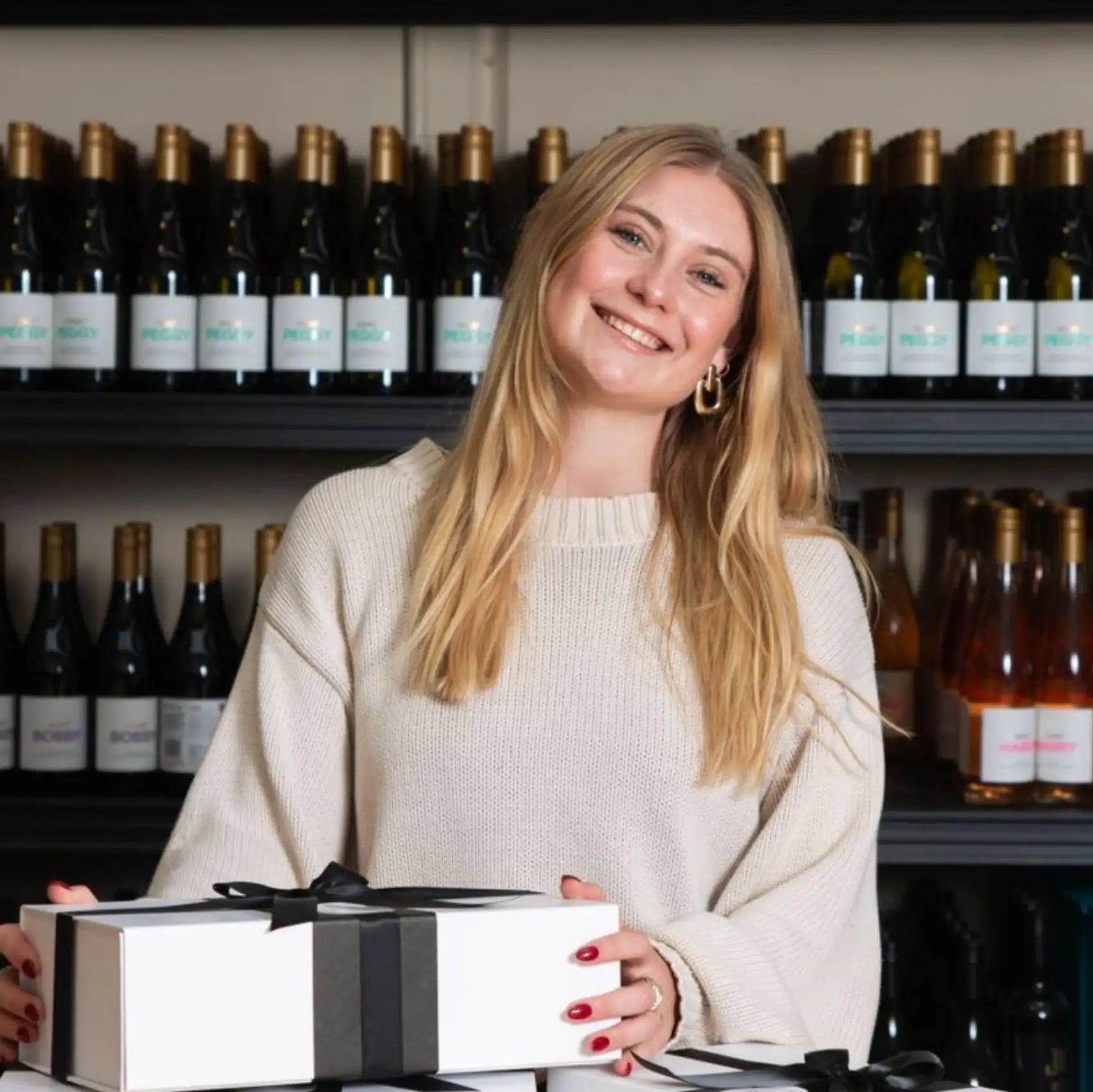 A woman in a cream-colored knit sweater holds a white gift box tied with black ribbon.