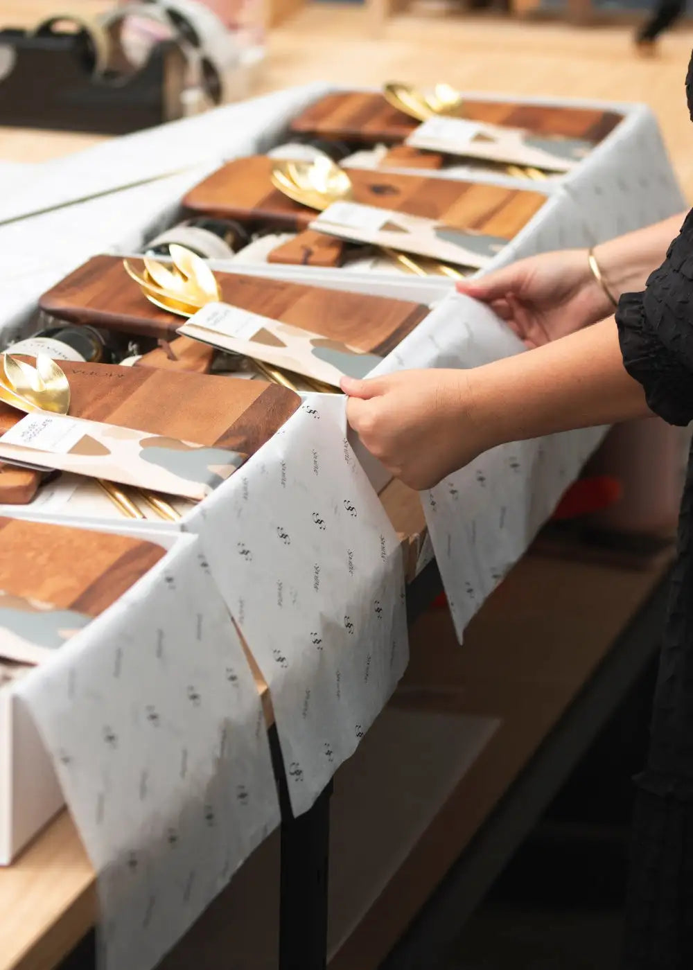 Wooden cutting boards with a natural grain finish, each accompanied by a gold-colored spoon and packaged in minimalist white paper.